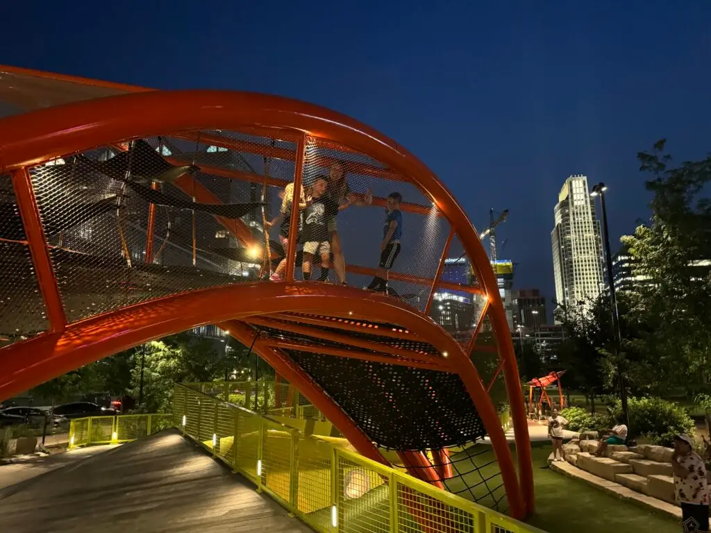 Children play on a brightly lit red playground structure at night, with an illuminated city skyline in the background.