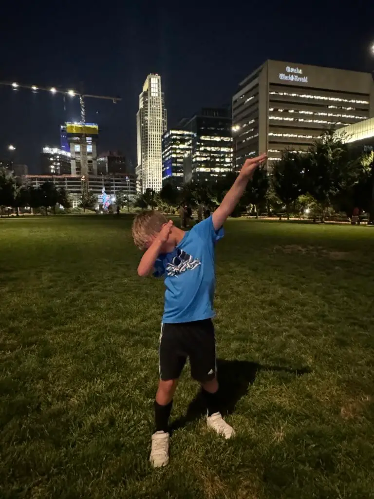 Child in a blue shirt and black shorts performs a dab pose on a grassy field at night, with illuminated buildings in the background.  Omaha with kids.