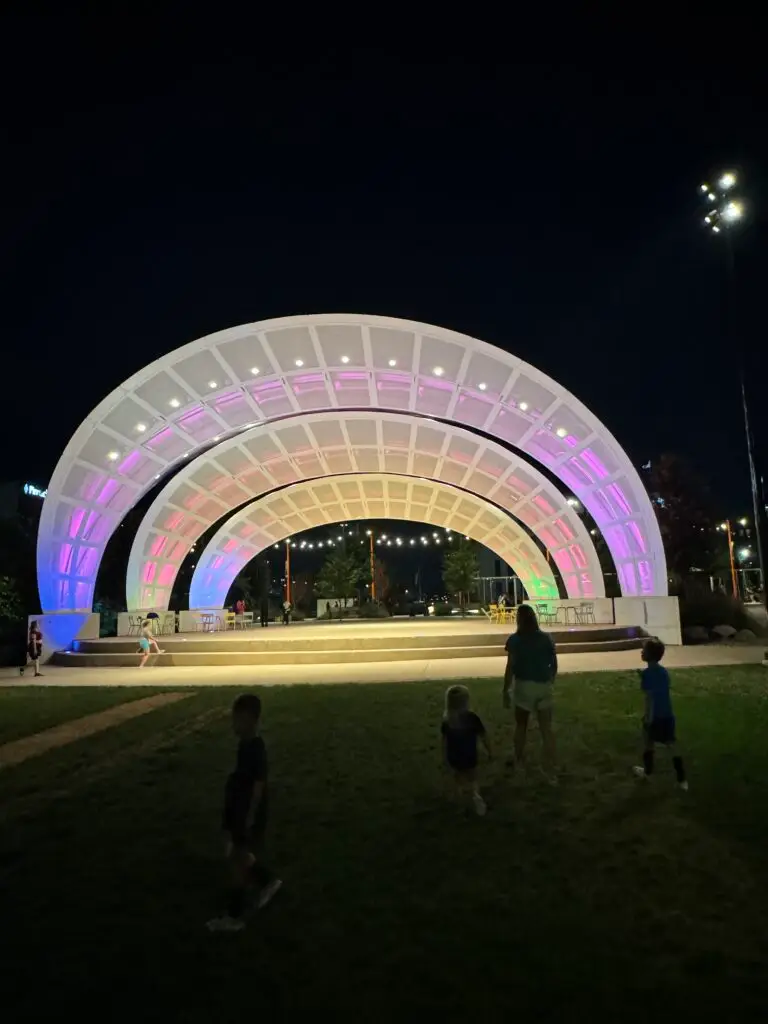 A person and children walk near a brightly lit, colorful amphitheater at night.   Omaha with kids.