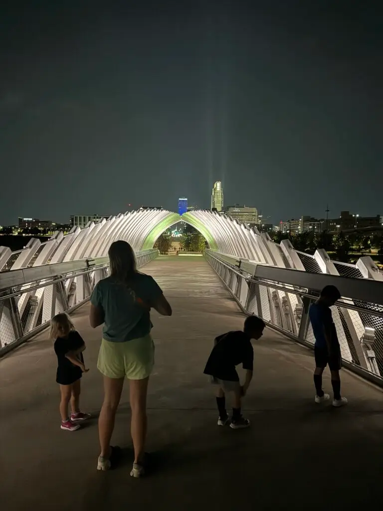 A woman and three children stand on a well-lit pedestrian bridge at night, with city buildings visible in the background.