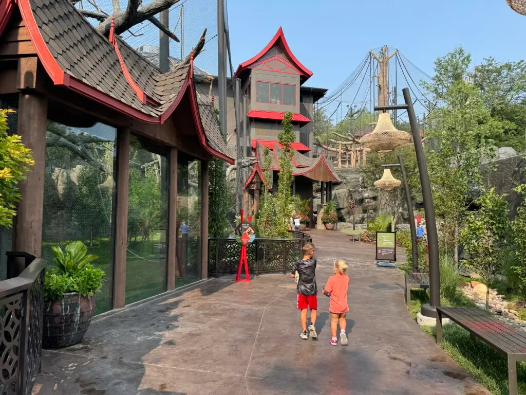 Two children walk on a zoo pathway with Asian-themed architecture and green vegetation on a sunny day.  Omaha Zoo with kids.