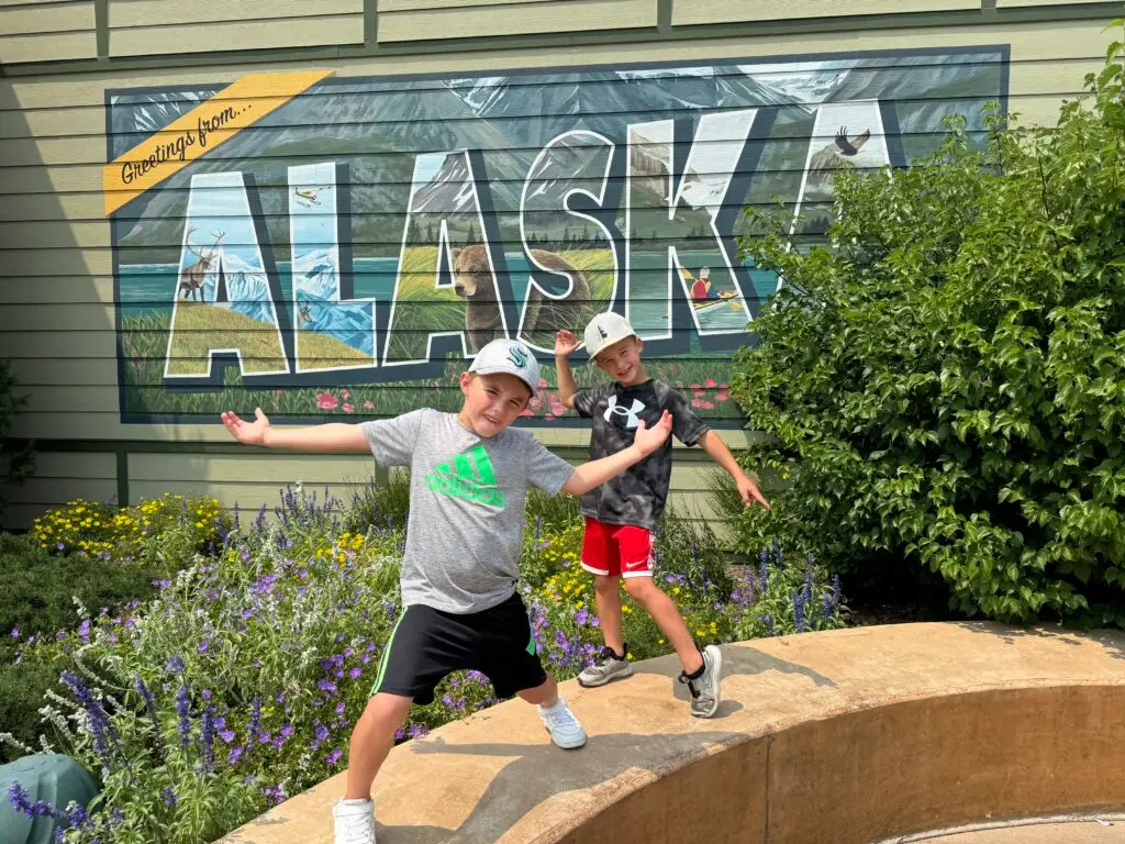 Two children pose energetically in front of an "Alaska" mural, surrounded by colorful flowers and greenery.  Omaha Zoo with kids.