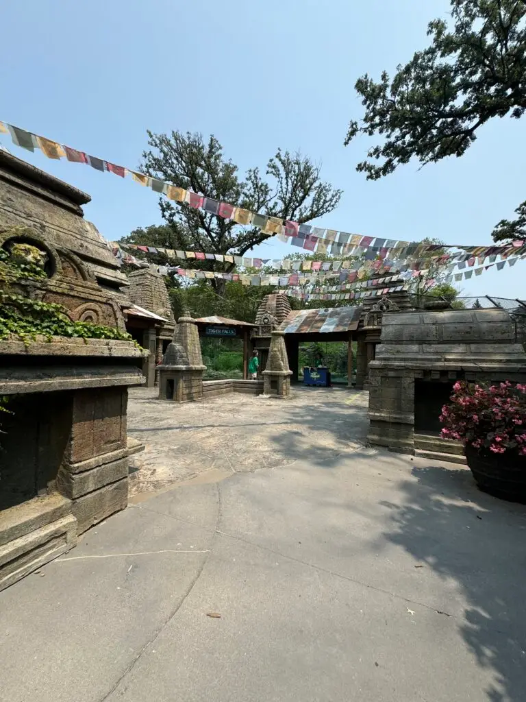 Outdoor temple-like structure with stone architecture, surrounded by trees and prayer flags hanging above.  Omaha Zoo with kids.