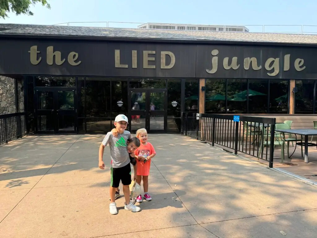 Three children stand in front of the entrance to the Lied Jungle building.  Omaha Zoo with kids.