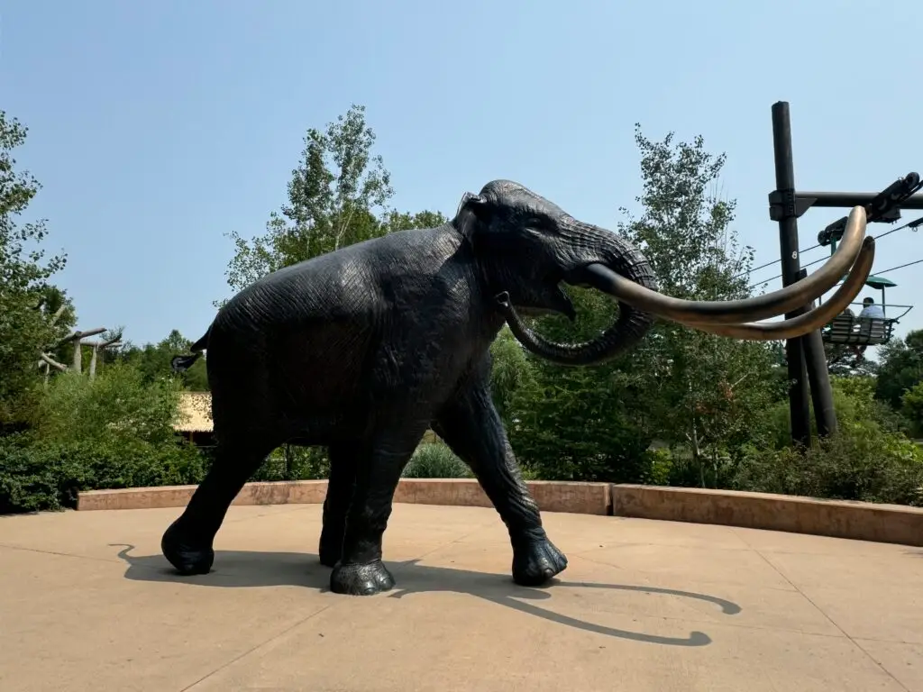 Bronze statue of a woolly mammoth stands on a concrete surface, surrounded by greenery and under a clear sky.  Omaha Zoo with kids.