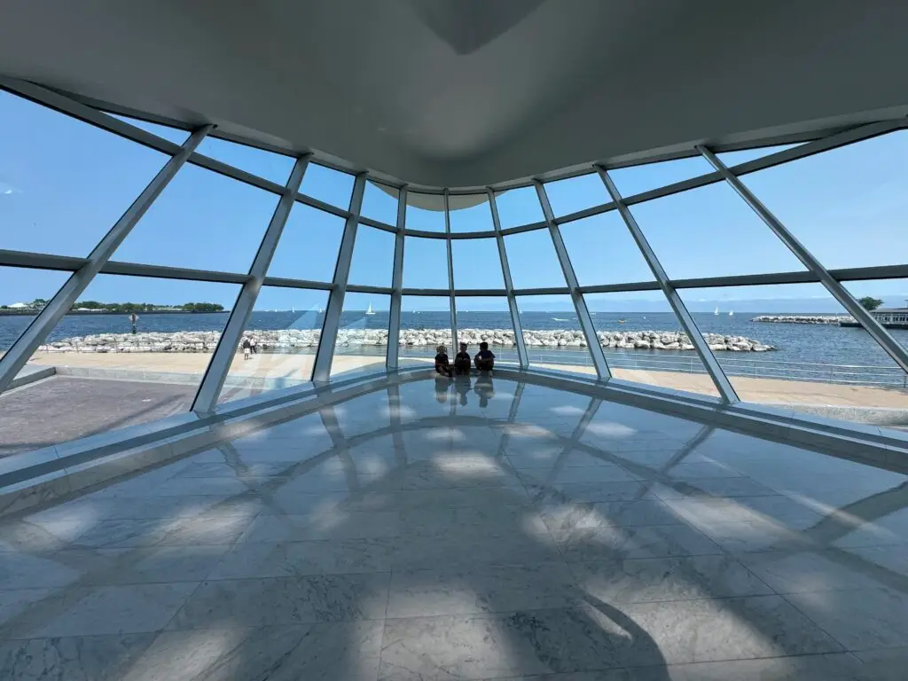 View of a waterfront through large, curved windows, with three people sitting on the floor, beneath a bright, clear sky.