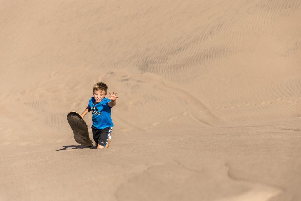 A child wearing a blue shirt slides down a sand dune on a sandboard, one hand reaching out.