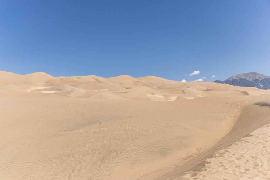 Expansive sand dunes under a clear blue sky, with distant mountains on the horizon.