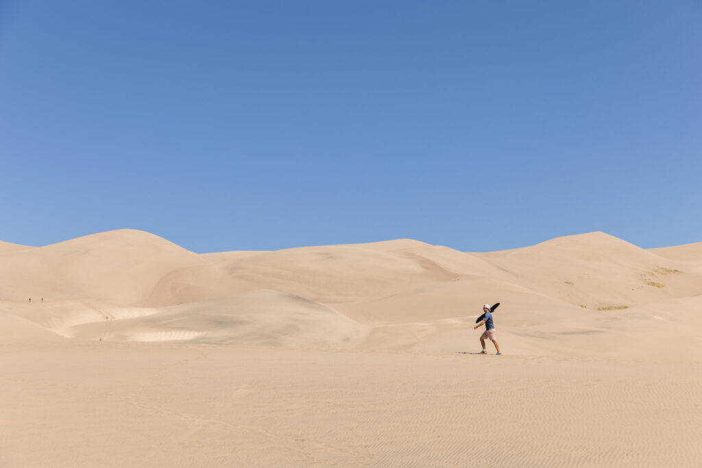 A person in a hat strolls across the vast Great Sand Dunes, navigating the rolling sand dunes beneath a clear blue sky—a perfect adventure for families with kids.