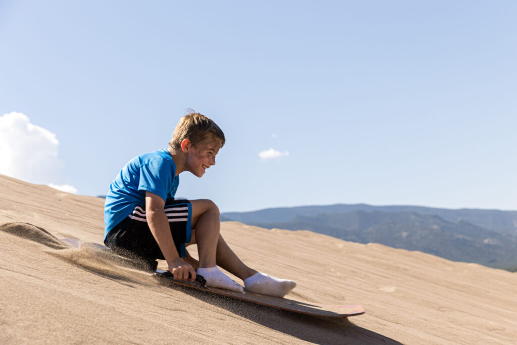 A boy sits on a sandboard, sliding down a dune at the Great Sand Dunes, wearing a blue shirt and dark shorts. The majestic mountains in the background frame this sunny adventure under a clear sky—perfect for kids seeking thrills in nature's vast playground.