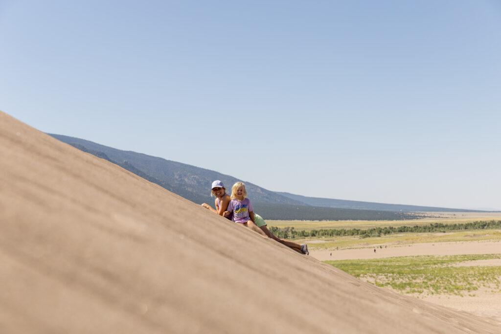 Two people sitting on a sloped sand dune under a clear blue sky, with mountains and greenery in the background.