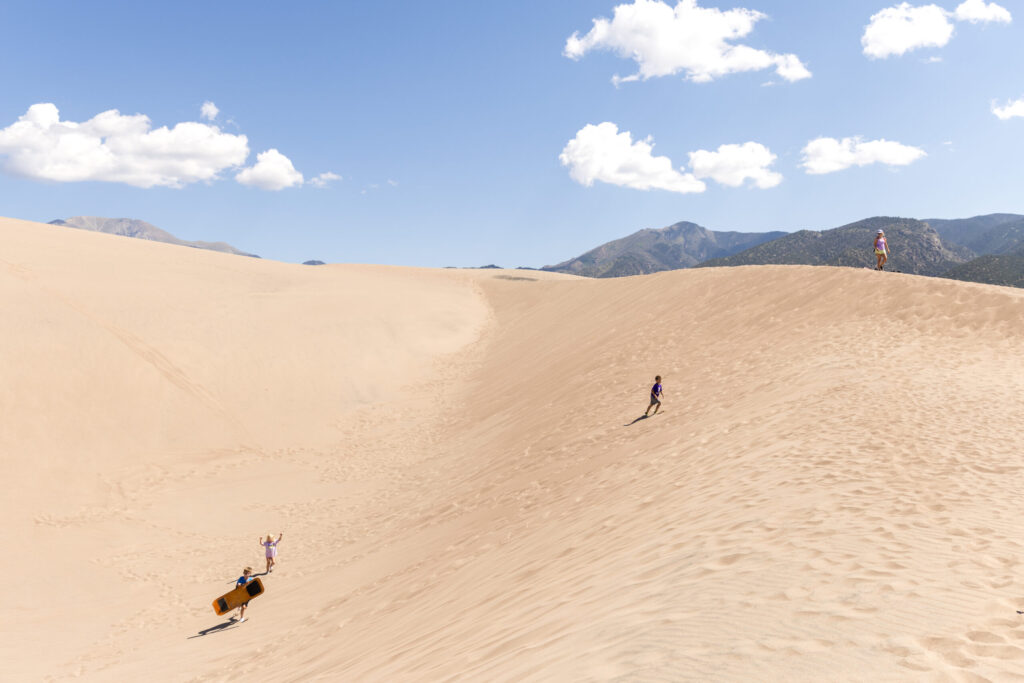 Three people explore the vast Great Sand Dunes under a clear blue sky, with majestic mountains in the background. One adventurous kid is sandboarding down a slope, leaving joyful trails in the golden sand.