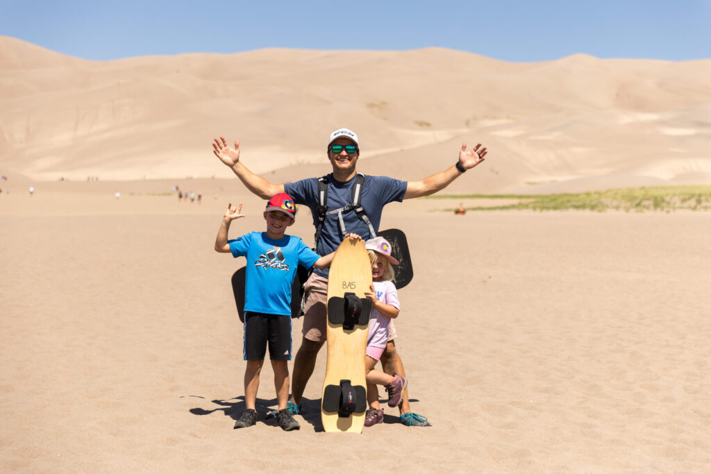 An adult and two children pose with a sandboard against the stunning backdrop of Great Sand Dunes, where the vast sandy landscape inspires adventure, and the adult's arms are raised in triumph.