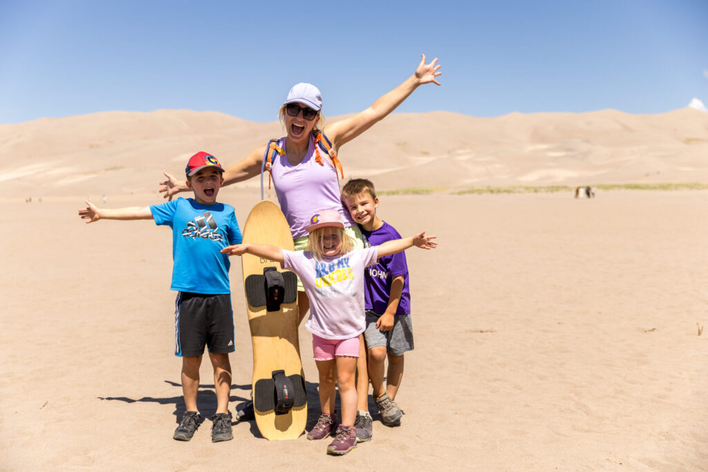 A smiling adult and three children stand with arms outstretched, posing with a sandboard against the stunning backdrop of the Great Sand Dunes, basking in the sunny desert landscape.