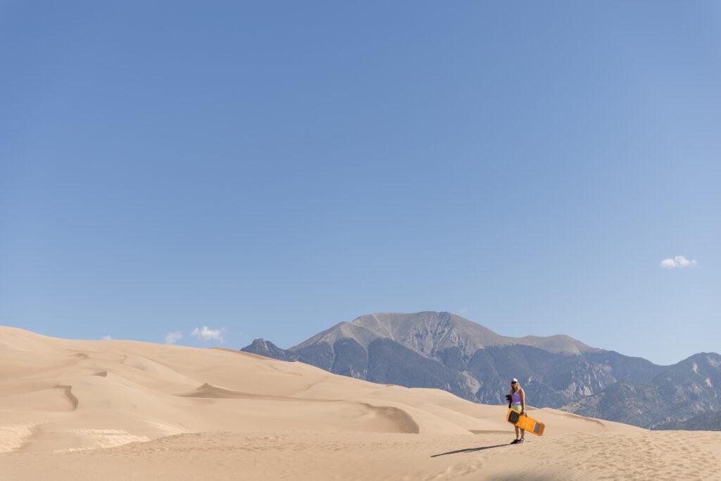 A person stands on sand dunes with a sandboard, in front of distant mountains under a clear blue sky.