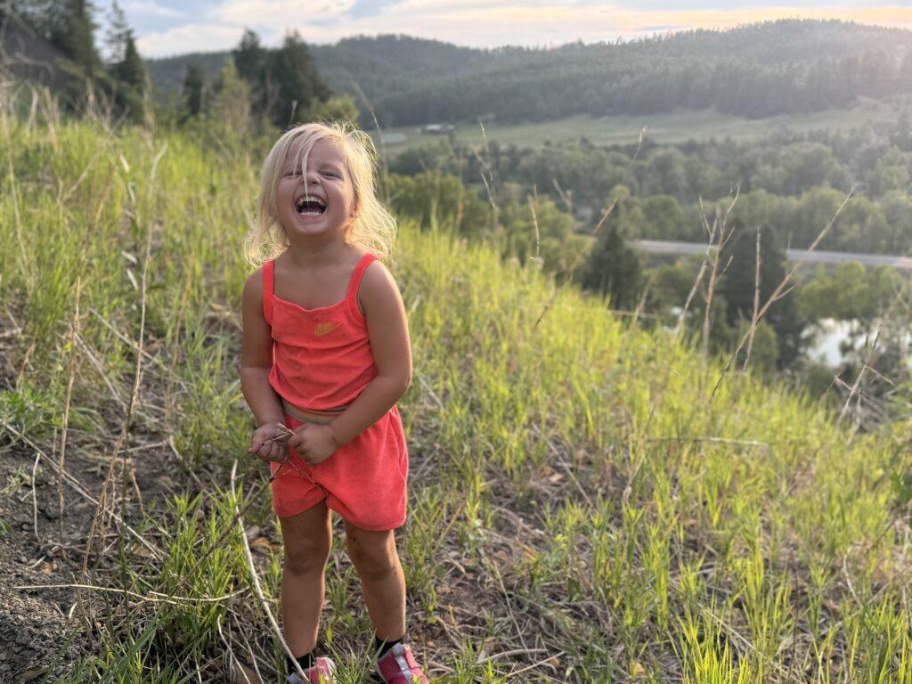 A young child in an orange outfit laughs while standing on a grassy hillside with a scenic view of trees and hills in the background.