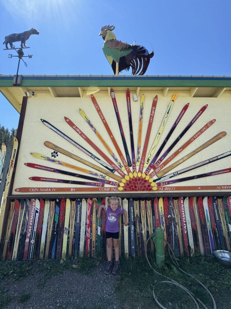 A child stands in front of a building in Pagosa Springs, decorated with skis arranged in a sunburst pattern, and topped with a whimsical rooster and bear weathervane.