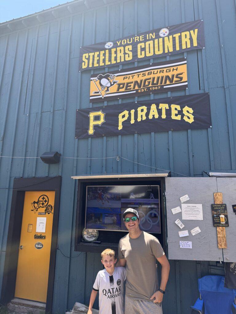 Man and boy smiling in front of a building adorned with Pittsburgh sports team signs: Steelers, Penguins, Pirates, sharing the same excitement as they did visiting Pagosa Springs with kids.