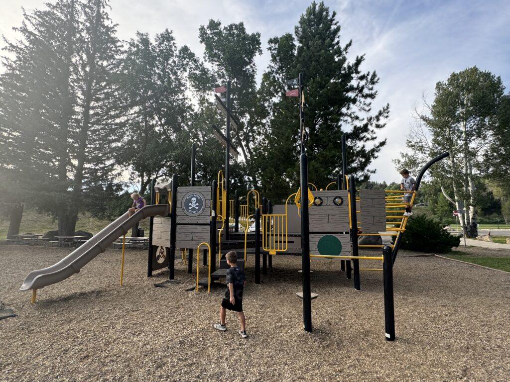 Children play on a playground structure designed like a pirate ship, featuring slides and climbing areas, surrounded by trees and a gravel ground.