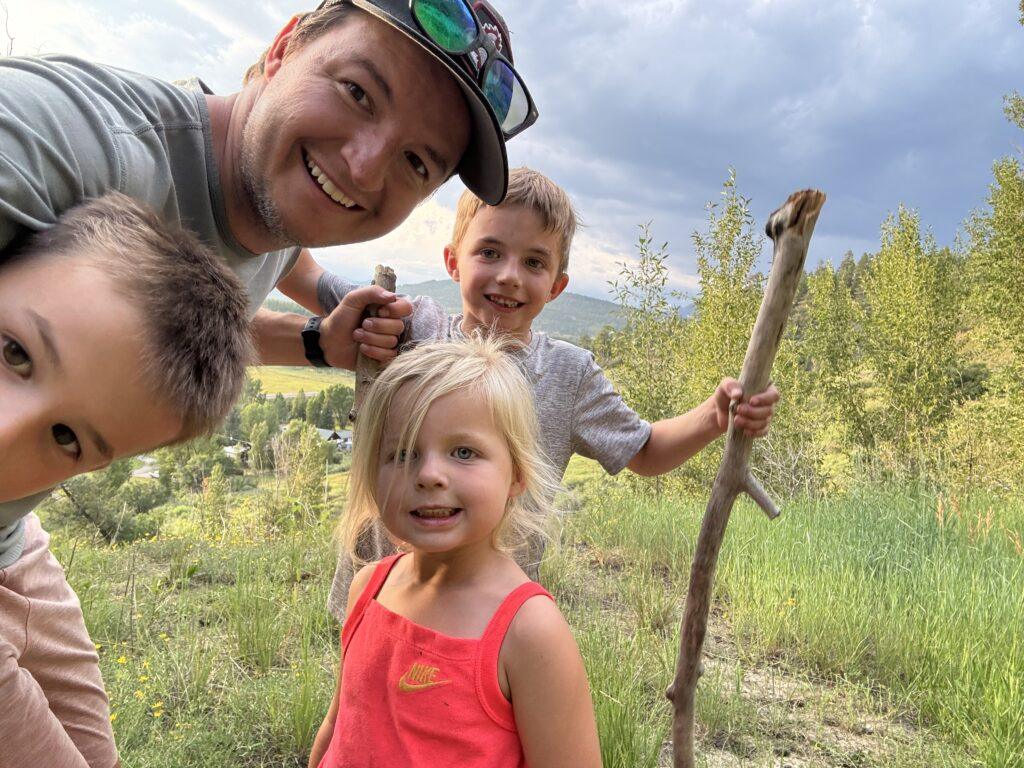 A man and three children pose outdoors on a grassy hill, surrounded by trees and under a cloudy sky. One child holds a long stick.