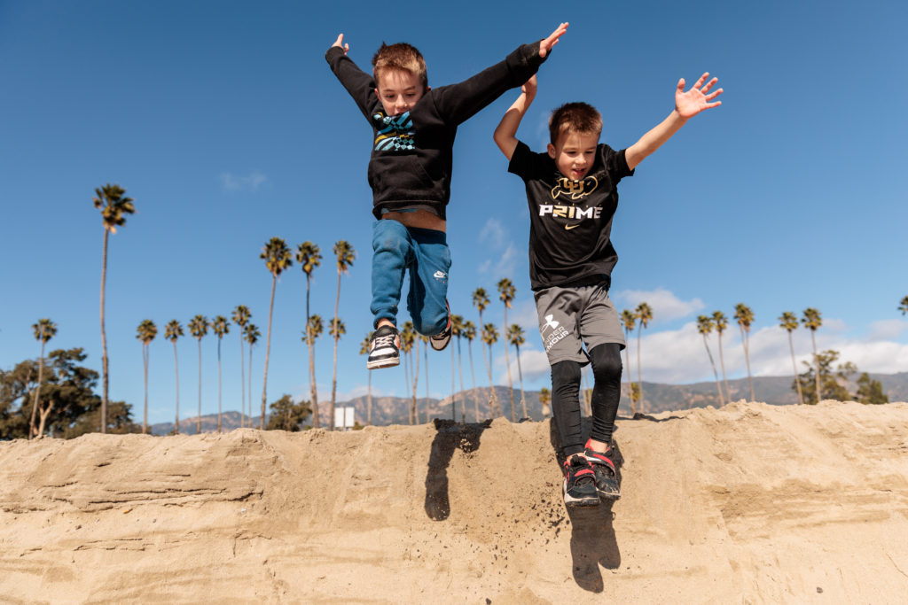 Two boys jumping over a sand pit with palm trees in the background.