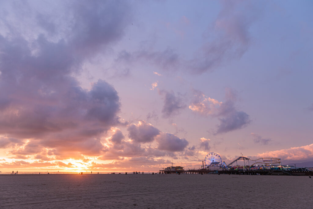 Sunset on a beach with a Ferris wheel and amusement park rides in the background, under a sky filled with clouds.