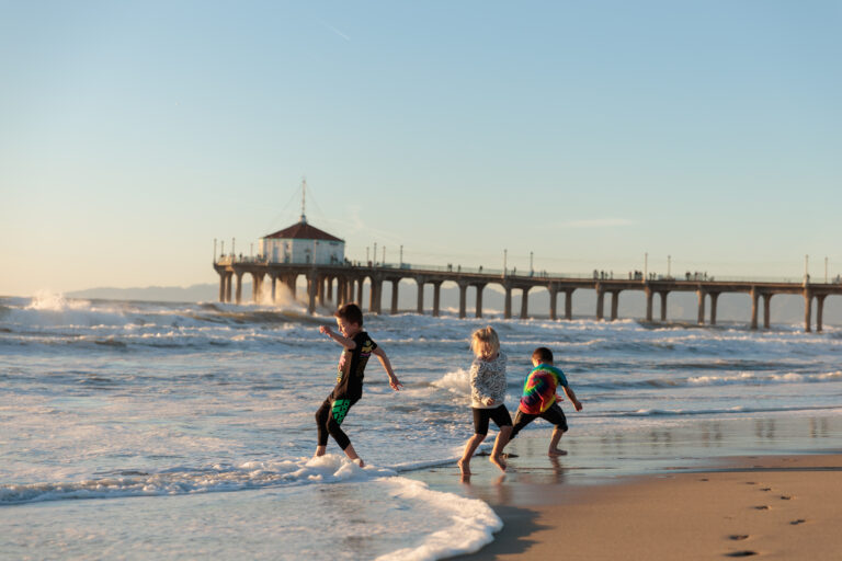 Three children play at the water's edge near a beach with a large pier in the background and gentle waves coming in. The sky is clear and the sun is setting, casting a warm glow over the scene. day in Manhattan Beach with kids