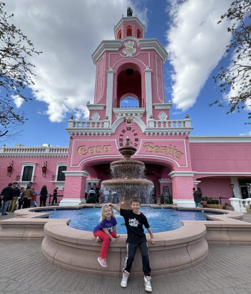 Two children pose in front of a large pink building labeled "Casa Bonita" with a fountain in the foreground and a clear blue sky above. Casa Bonita with the family.