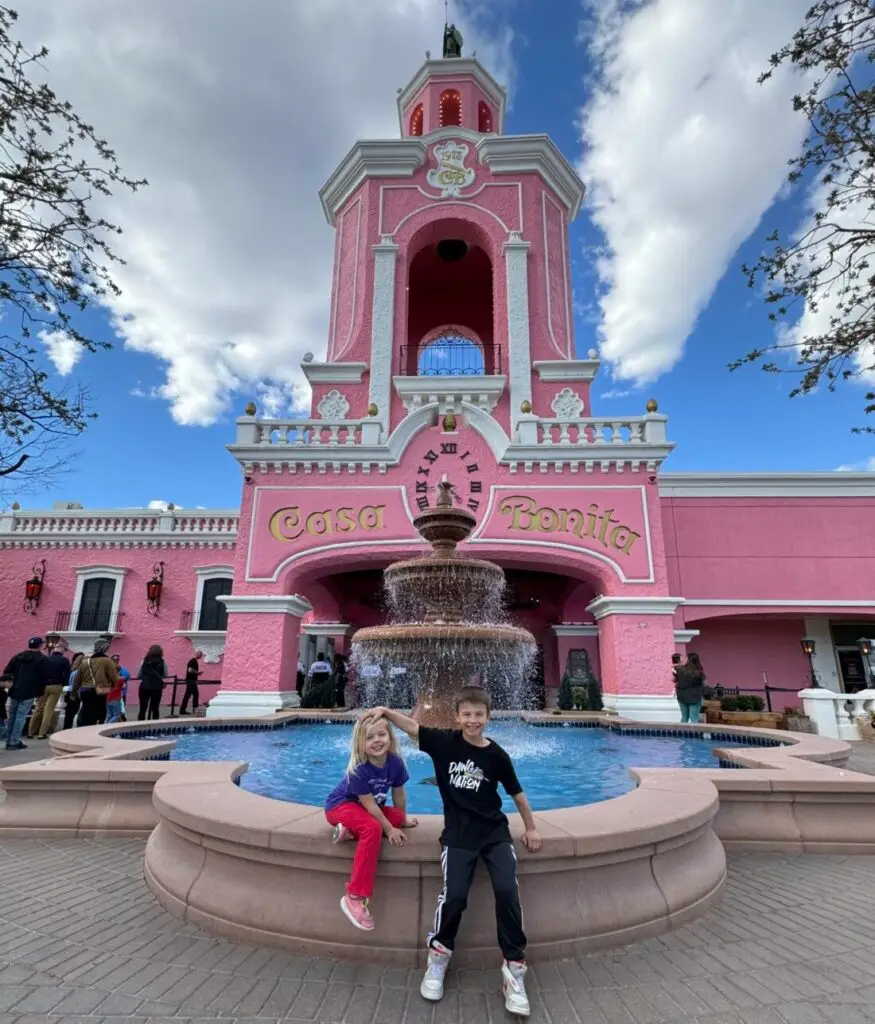 Two children pose in front of a large pink building labeled "Casa Bonita" with a fountain in the foreground and a clear blue sky above. Casa Bonita with the family.