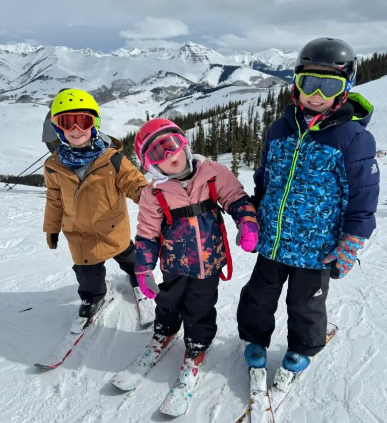 Three children in winter clothing and ski gear are standing on a snowy slope with a mountainous background. They are equipped with skis, helmets, and goggles, and two of them have ski poles. Crested Butte Spring Break