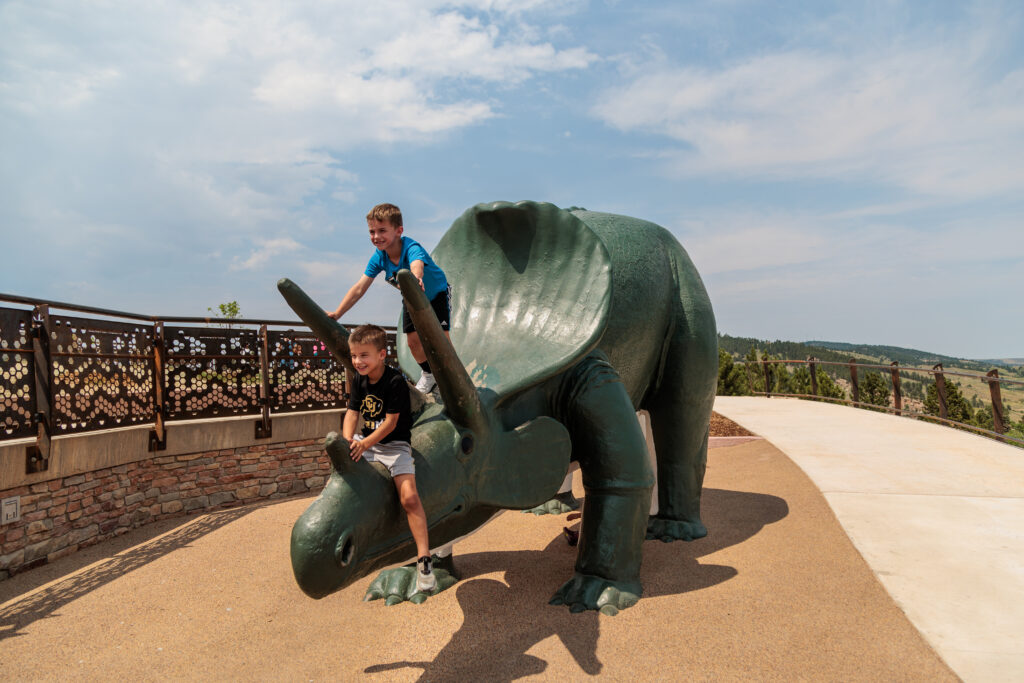 Two children are playing on a large triceratops statue outdoors. One child is sitting on top of the head, and the other is sitting on the neck. A walkway and railing are visible in the background.  Free things to do in Rapid City with kids.