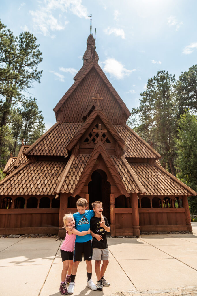 Three children stand in front of a wooden church with intricate carvings, set against a backdrop of tall trees and a partly cloudy sky.  Free things to do in Rapid City with kids.