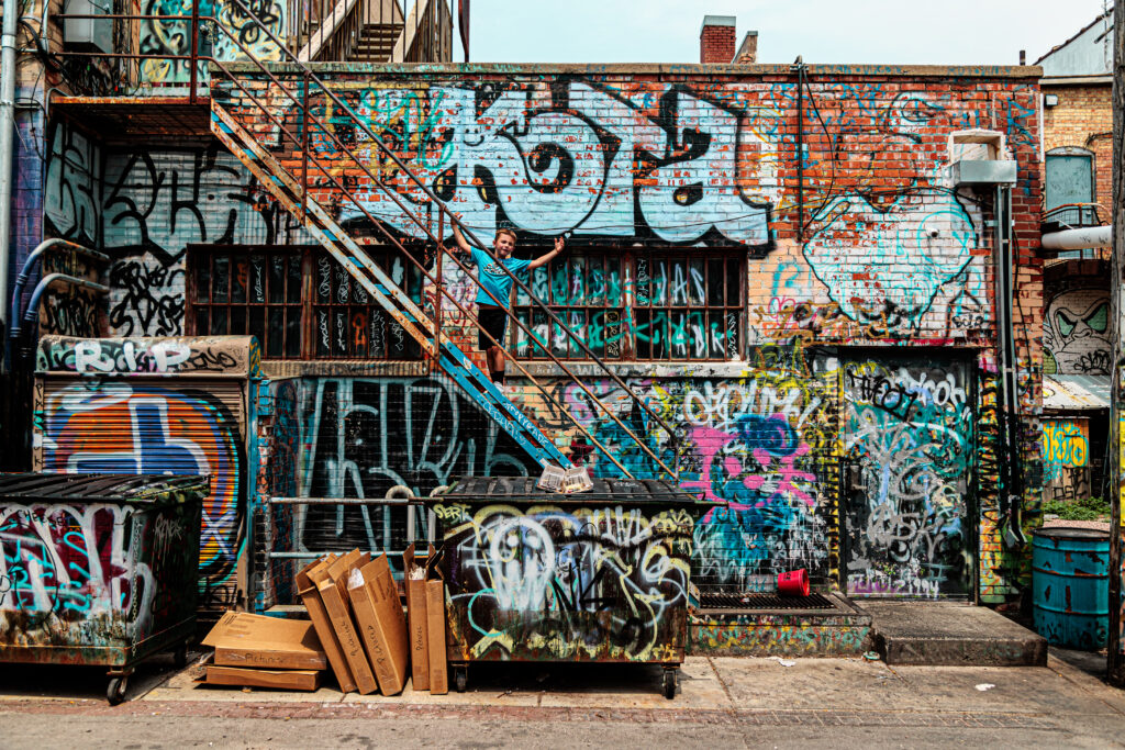 A person stands on a metal staircase against a heavily graffiti-covered brick wall, with dumpsters and cardboard boxes visible on the ground below. Free things to do in Rapid City with kids.