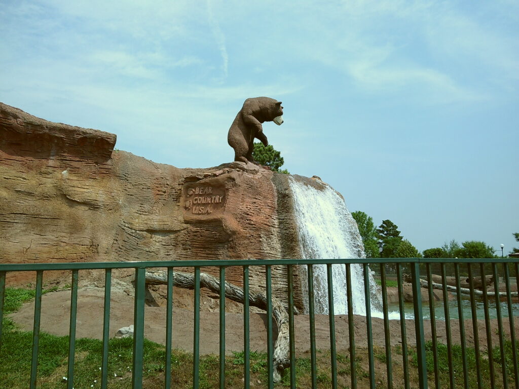 Statue of a bear standing on a rock near a waterfall, with the words "Bear Country USA" carved into the rock, surrounded by a fence. South Dakota with Kids, Bear Country USA.