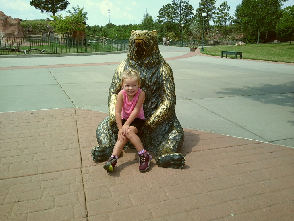 A young girl in a pink top and black shorts sits on the lap of a large bear statue in a park area with trees and pathways in the background.  South Dakota with Kids, Bear Country USA.