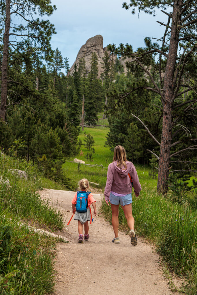 An adult and a child with a blue backpack walk on a dirt trail through a forested area, heading towards a large rock formation in the distance.  Custer State Park with kids.