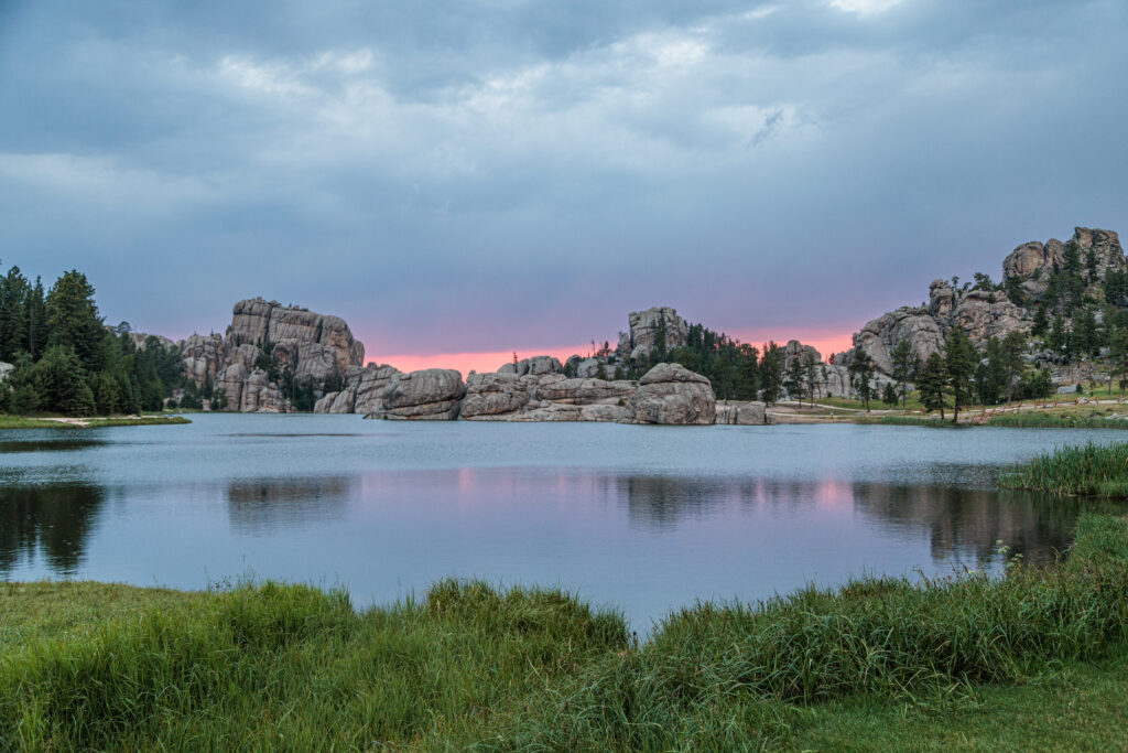 A serene lake with large rock formations and trees on the shore, under a cloudy sky with a pink glow from the setting sun. Custer State Park with kids. Custer State Park with kids.