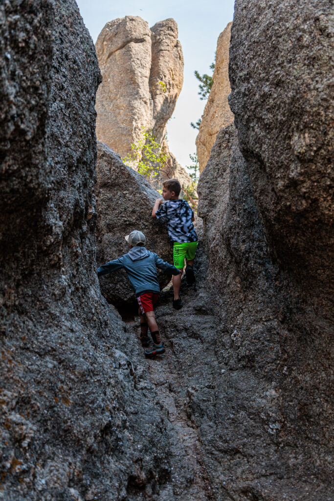 Two children climb between large rock formations in a narrow passage, with one child wearing a grey jacket and red shorts and the other in a black and white shirt with neon green shorts. Custer State Park with kids.