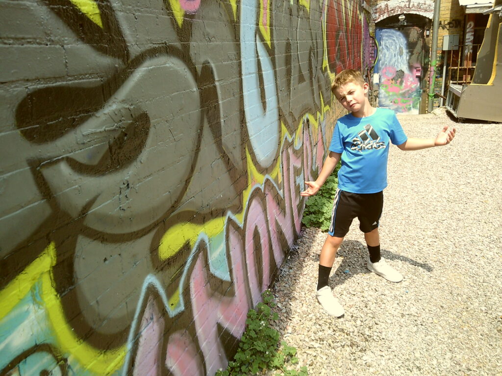 Child in a blue shirt posing confidently in front of a colorful graffiti-covered wall on a gravel path. Camp Snap Discount Code