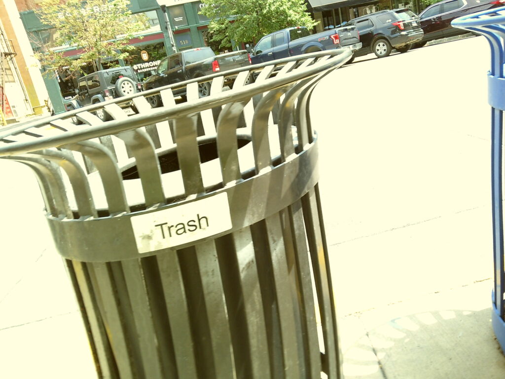 A black trash can on a sidewalk with cars and trees in the background.  Camp Snap Discount Code