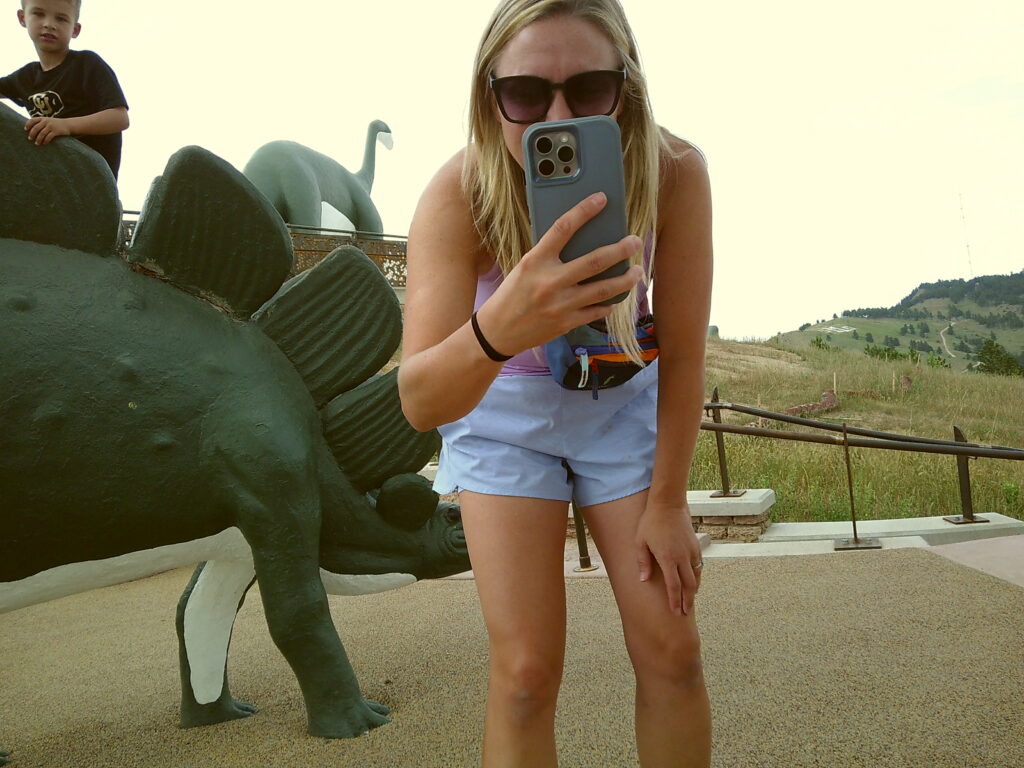 A person taking a photo with a smartphone stands next to a dinosaur sculpture in an outdoor setting. A child is in the background. Camp Snap Discount Code