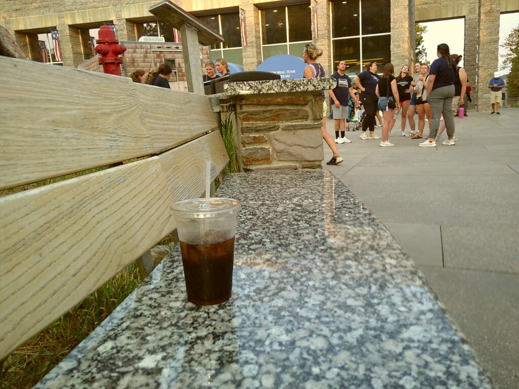 A plastic cup with a straw sits on a stone bench near a wooden fence. People gather in the background near a building and a red fire hydrant. Camp Snap Discount Code