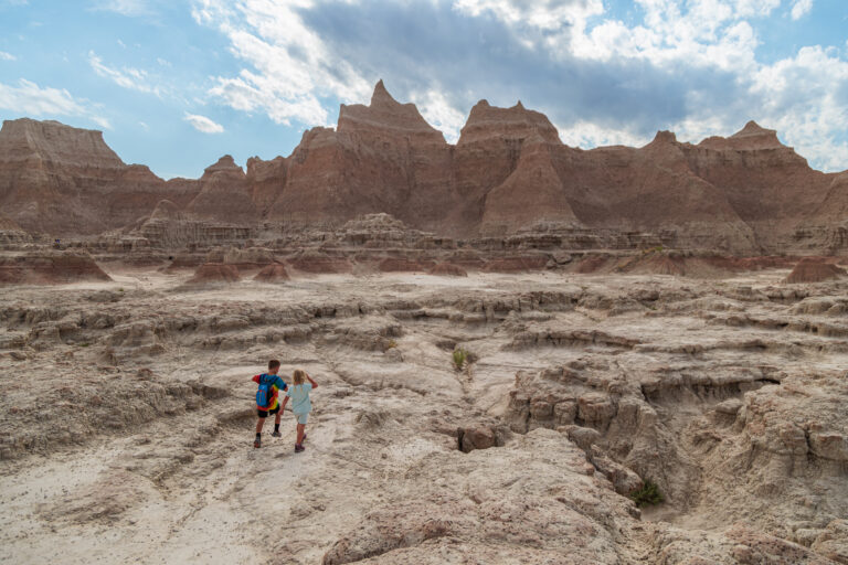 Two children walk through a rocky, arid landscape with rugged hills and a cloudy sky in the background. Badlands National Park