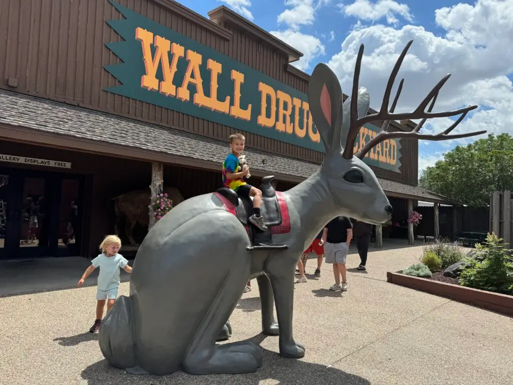 Two children interact with a large jackalope statue outside Wall Drug. One sits on the statue while the other stands beside it. Wall Drug with kids