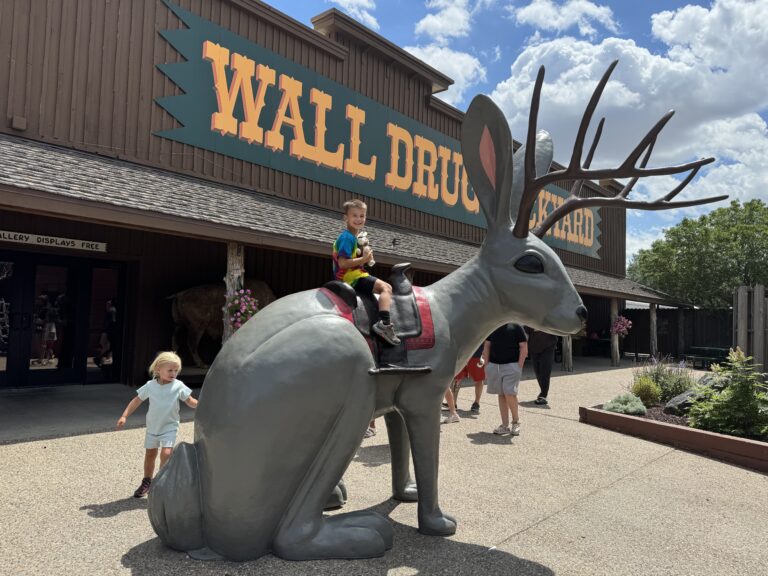 Two children interact with a large jackalope statue outside Wall Drug. One sits on the statue while the other stands beside it. Wall Drug with kids