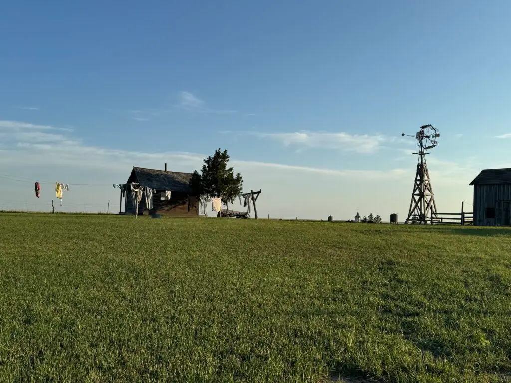 A rural scene with a wooden house, clothesline, windmill, and barn set against a clear blue sky and green grass. 1880 Town with kids