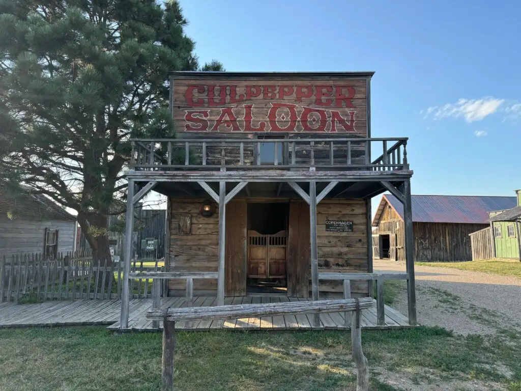 Old Western-style wooden saloon with "Culpepper Saloon" sign, situated in a rustic setting with clear blue sky. 1880 Town with kids