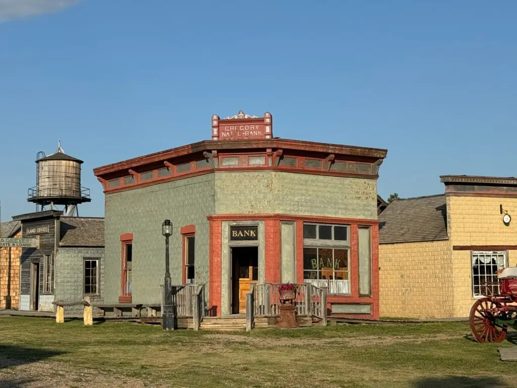 Old-fashioned bank building with a water tower nearby, set in a rustic, historical town. Red and green paint adorns the bank, with wooden structures surrounding the area.