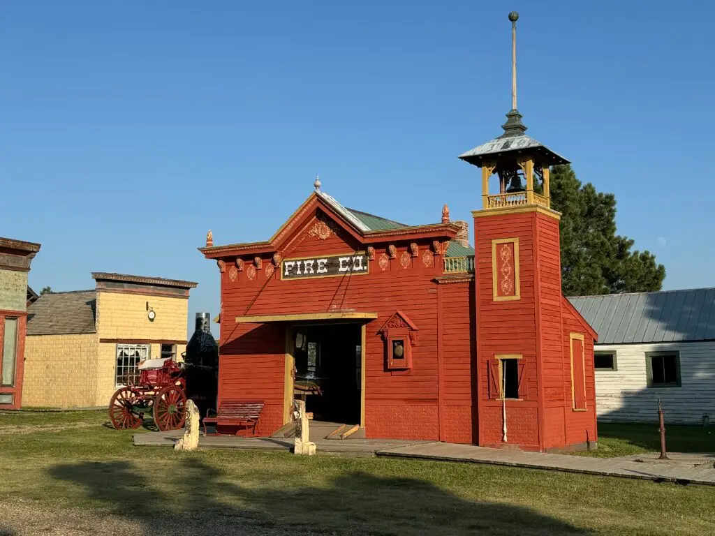 A vintage fire station with a red facade and a bell tower, set in a grassy area, under a clear blue sky. An old-fashioned fire engine is parked beside it. 1880 Town with kids