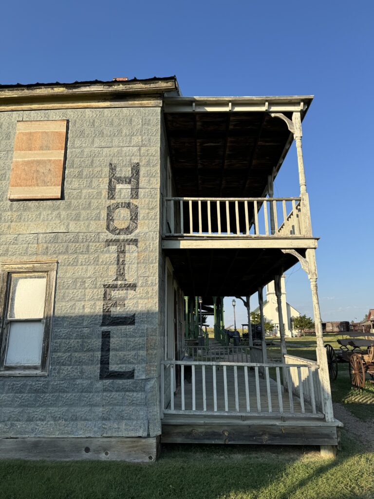 Side view of an old, weathered wooden building with a faded "Hotel" sign. The building has a covered porch and boarded-up windows, set against a clear blue sky. 1880 Town with kids